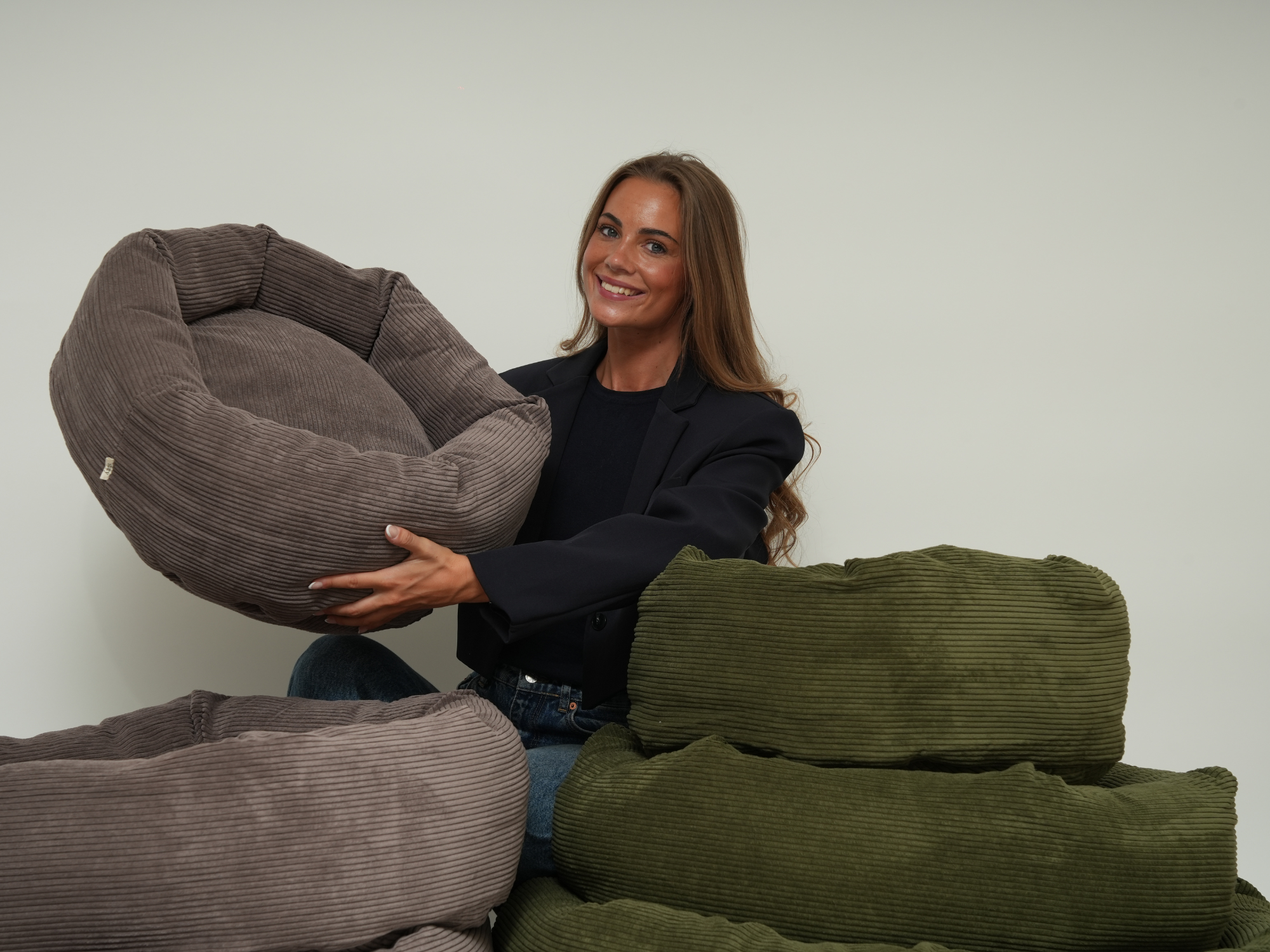 Woman holding a taupe corduroy dog bed beside a stack of moss green beds